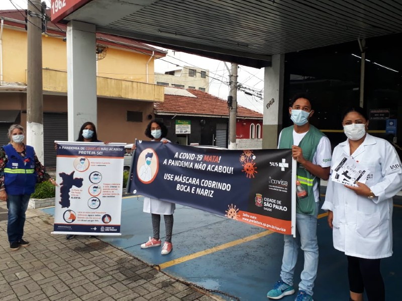 Foto da psicóloga Valéria Chagas durante uma ação de conscientização contra a Covid-19. Ela está com outras quatro pessoas segurando faixas e cartazes com recomendações para evitar a contaminação. Todos usam máscara e estão respeitando o distanciamento social